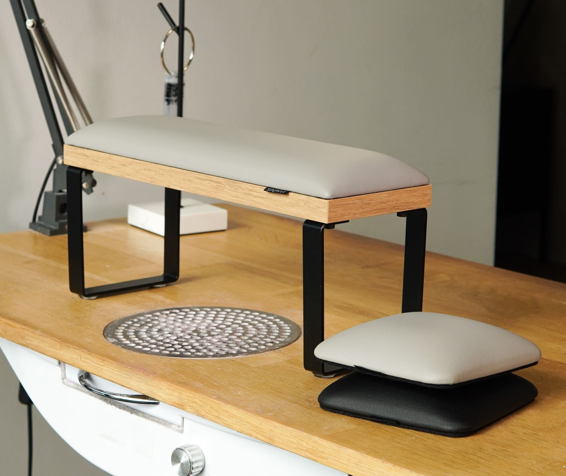 SPENVI Loft manicure hand rest with light grey cushion and black metal legs on a wooden salon desk. Includes matching SPENVI elbow rest cushion for ergonomic nail technician comfort in modern salons.