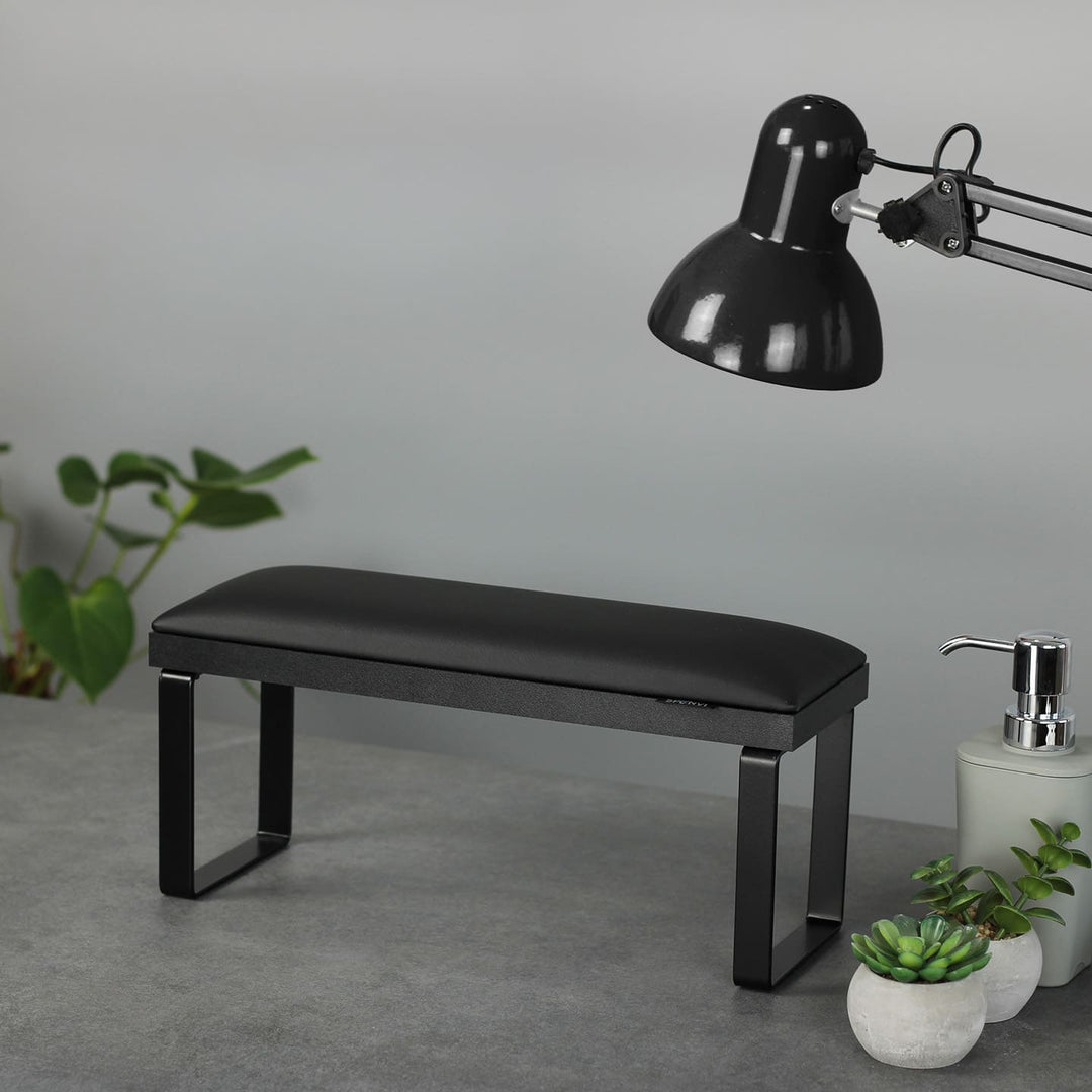 SPENVI Loft manicure arm rest in black with black leather cushion, black metal legs, and black base on a salon desk with plants and lamp.