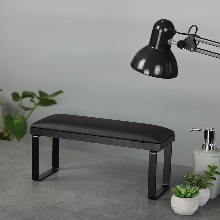 SPENVI Loft manicure arm rest in black with black leather cushion, black metal legs, and black base on a salon desk with plants and lamp.